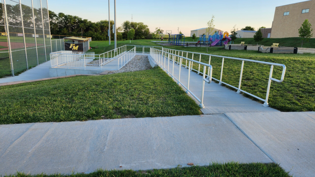 Outdoor school ramp with white railings leading to a playground and sports field.