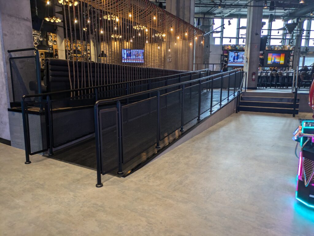 Black perforated metal panel handrail installed along a ramp at a restaurant bar entrance with hanging lights above.